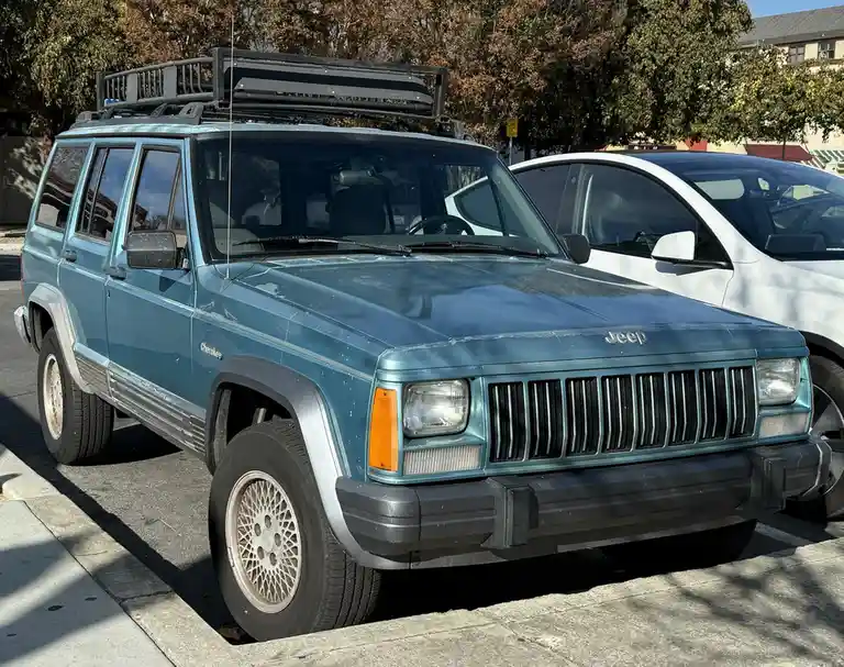 A light blue 1990s Jeep Cherokee SUV is parked in a lot, featuring a black roof rack and silver trim, with a white SUV…