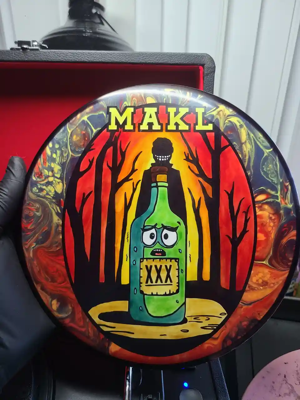 A hand in a black glove holds a custom-painted disc golf disc. The disc features a cartoonish green bottle with a terrified…