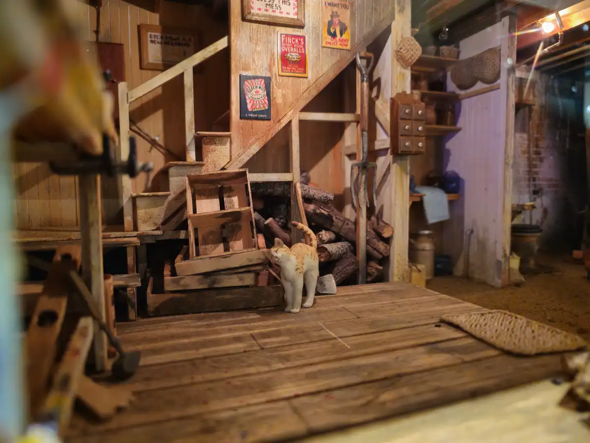 A miniature diorama of a rustic, cluttered wooden cabin interior with a small figurine cat standing near stacked crates and…