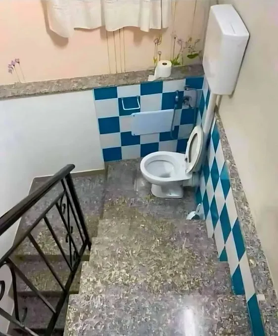 a white toilet is installed on a narrow staircase with a black metal railing, surrounded by blue and white checkered tiles…