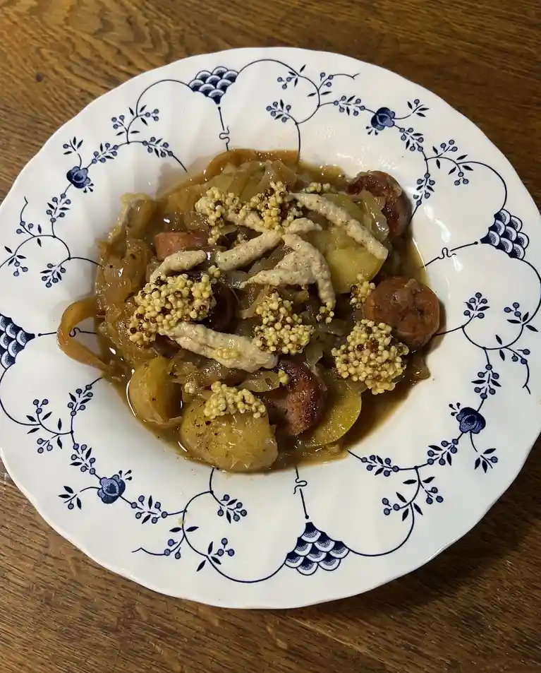 a close-up photo of a white ceramic plate with blue floral patterns, holding a dish of sauerkraut, small sausages, potatoes…