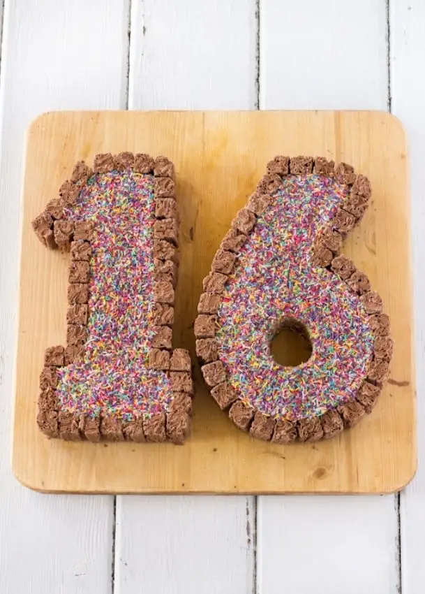 A cake shaped like the number 16, made from chocolate cereal pieces and covered in colorful sprinkles, sits on a wooden…