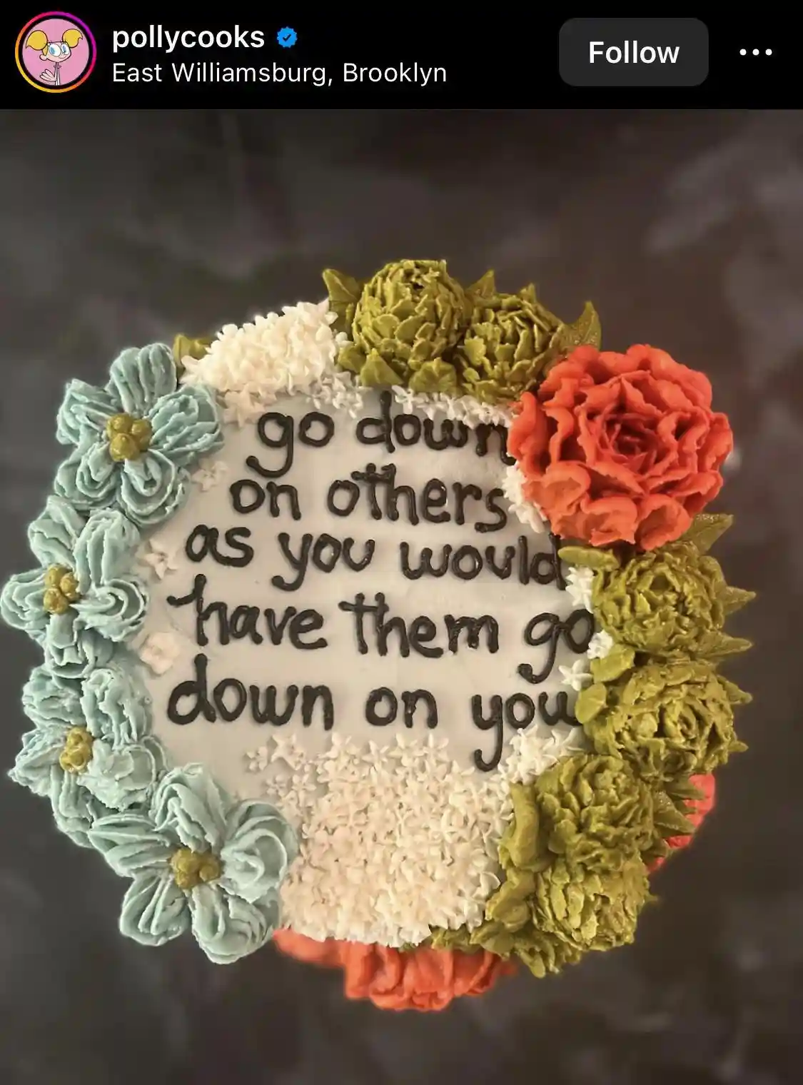 A round white cake decorated with piped frosting flowers in teal, white, olive green, and coral red, arranged in a border…