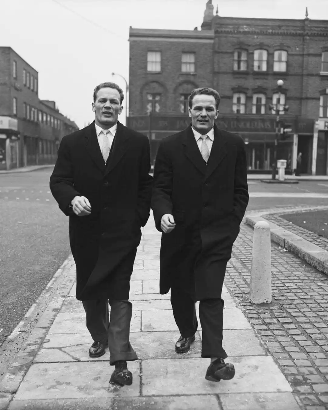 Two men in dark overcoats and ties walk down a city sidewalk, their faces showing mild surprise or concern. Their shoes are…