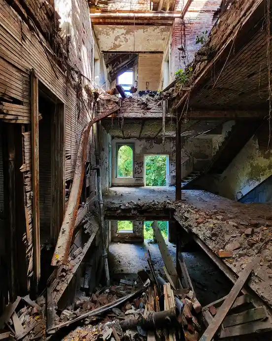 An image of a severely dilapidated, multi-level abandoned building with collapsed floors, exposed brick, rusted metal beams…