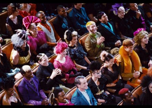 An audience of people in colorful, theatrical costumes sits in rows, many looking emotional or tearful. The overlaid text…