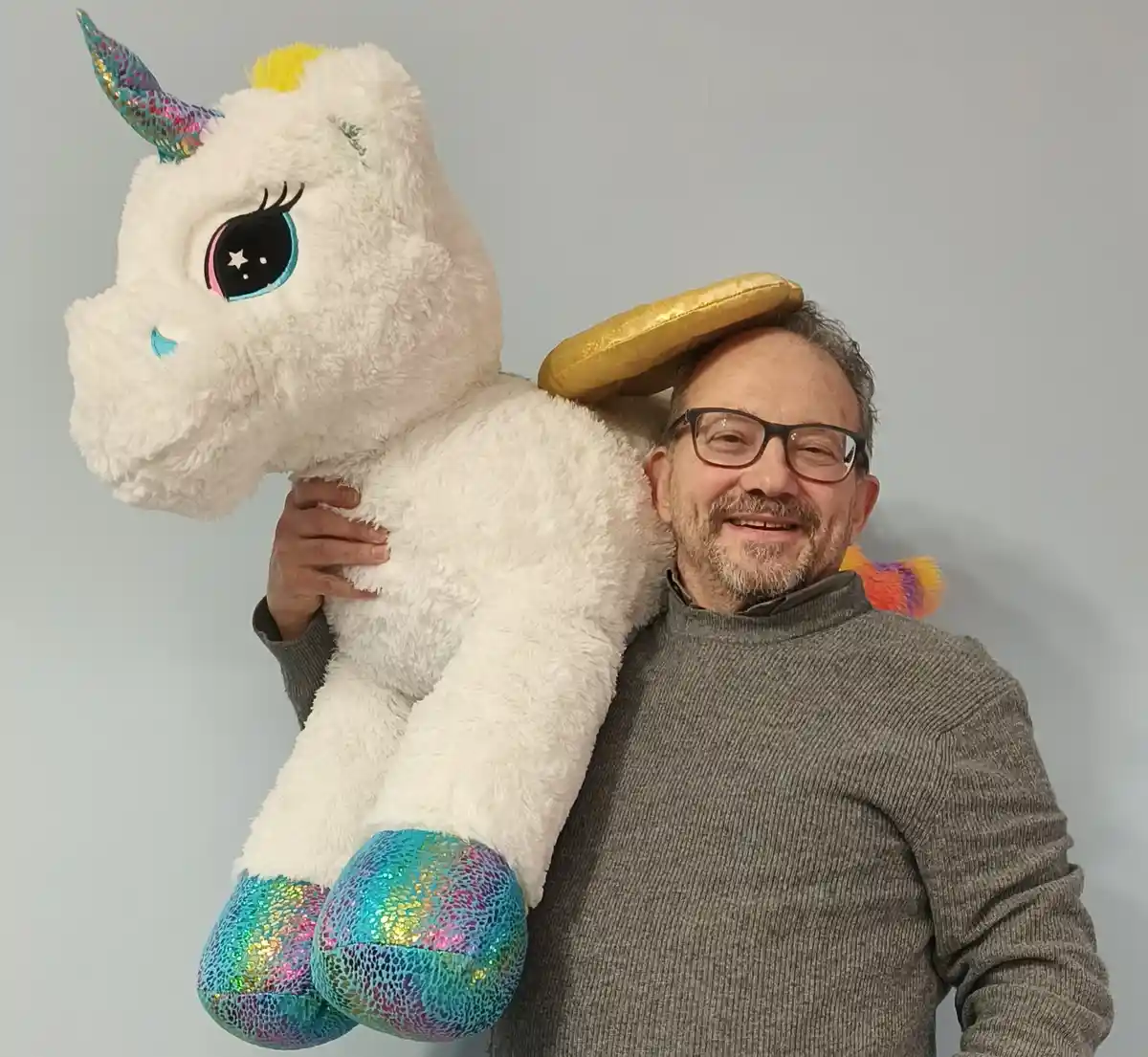 A smiling man with glasses and a grey sweater holds a large white plush unicorn with glittery rainbow hooves and horn. A…