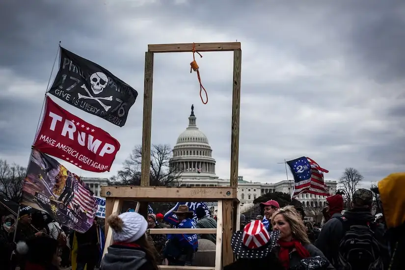 A crowd of protesters holding Trump flags and a pirate flag stands before a wooden gallows with a noose hanging from it…