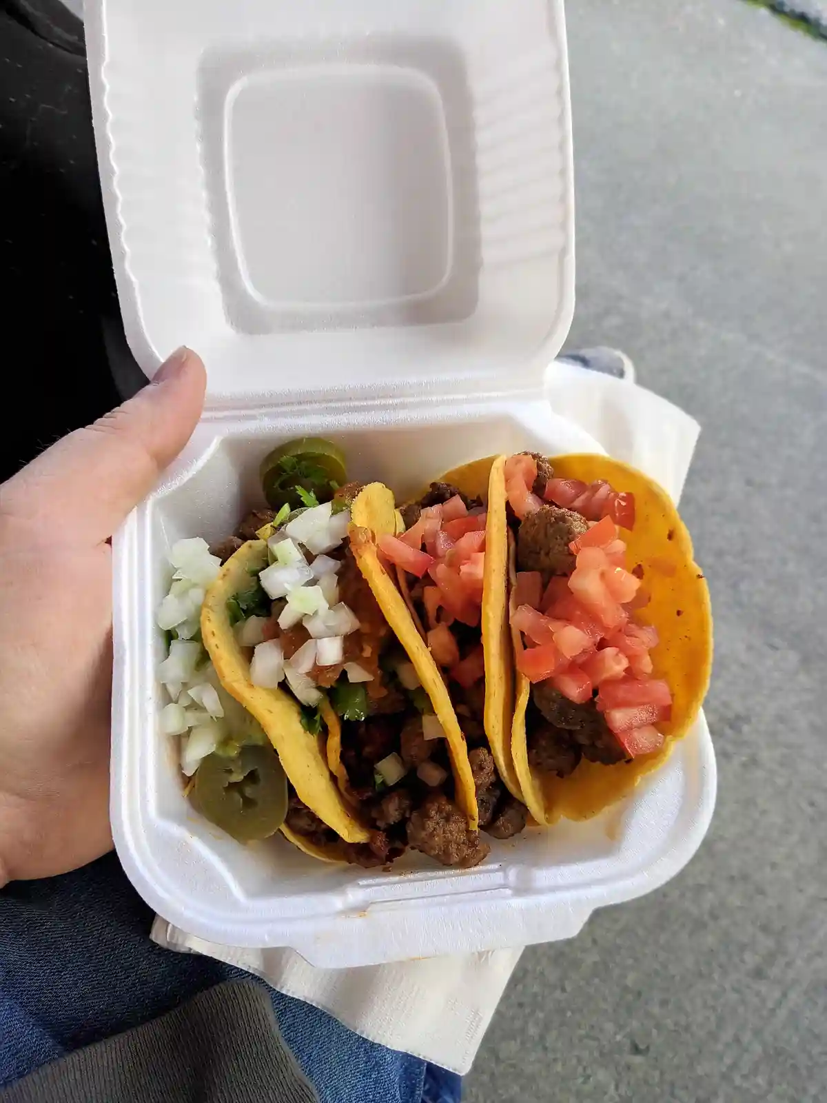 A hand holds an open white styrofoam takeout container filled with three soft tacos topped with meat, diced tomatoes…