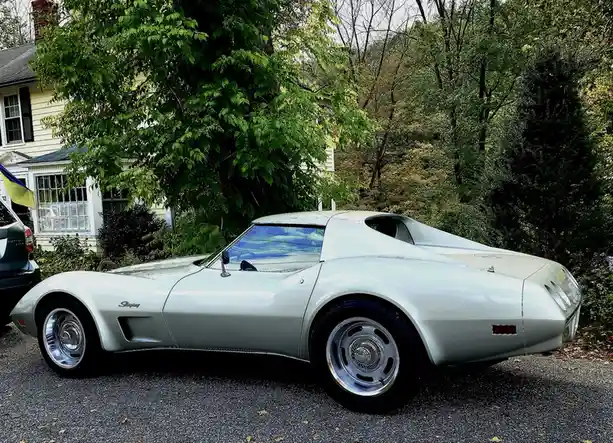 A silver 1970s Chevrolet Corvette Stingray is parked on a gravel driveway in front of a house with green trees. The car has…