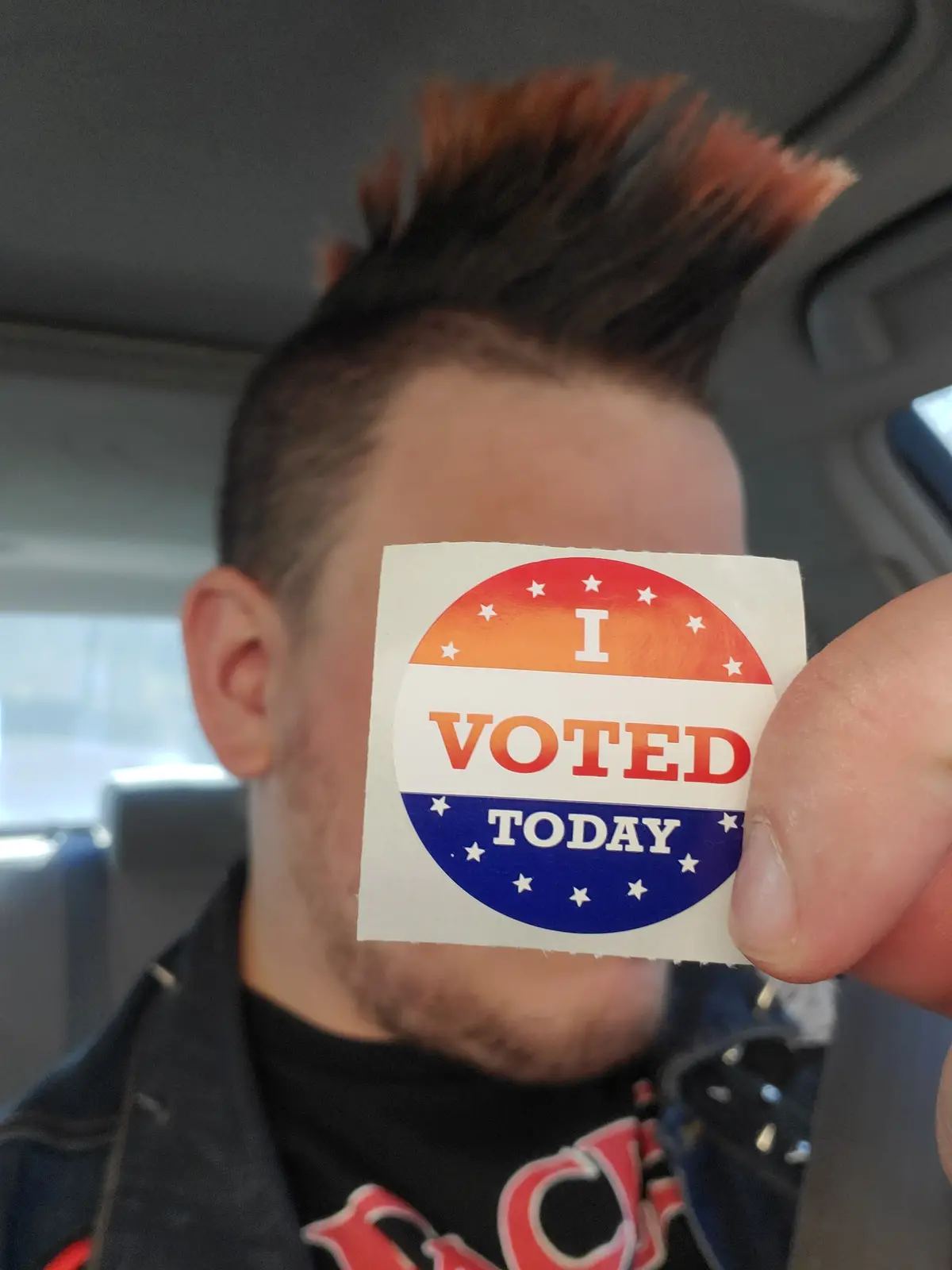 A person with a mohawk hairstyle is holding up a circular 'I VOTED TODAY' sticker in front of their face, obscuring it…