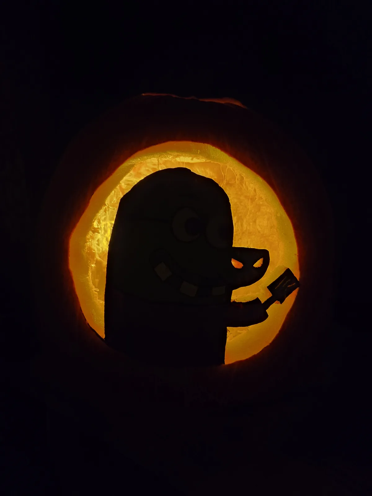 A carved pumpkin with a glowing orange interior features a silhouette of a Minion character holding a small tool, with the…