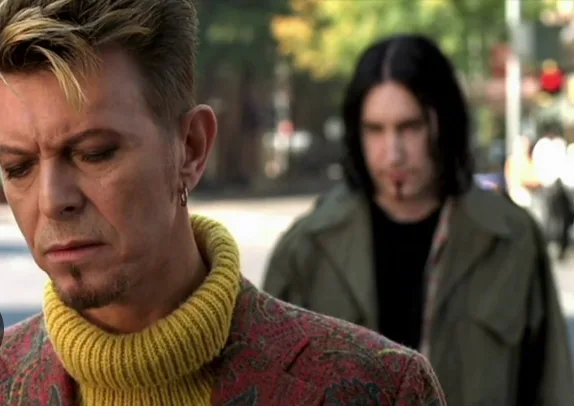 A man with blonde hair and a yellow turtleneck looks down with a displeased expression, while another man with long dark…