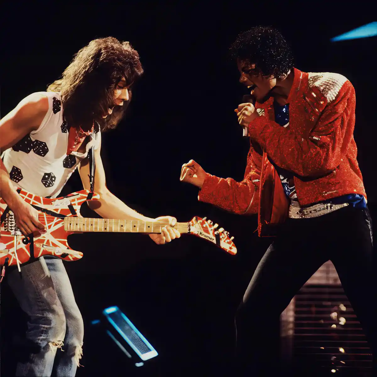 A photo of two musicians on stage, one holding a guitar with dice-patterned clothing, the other in a red sparkly jacket…