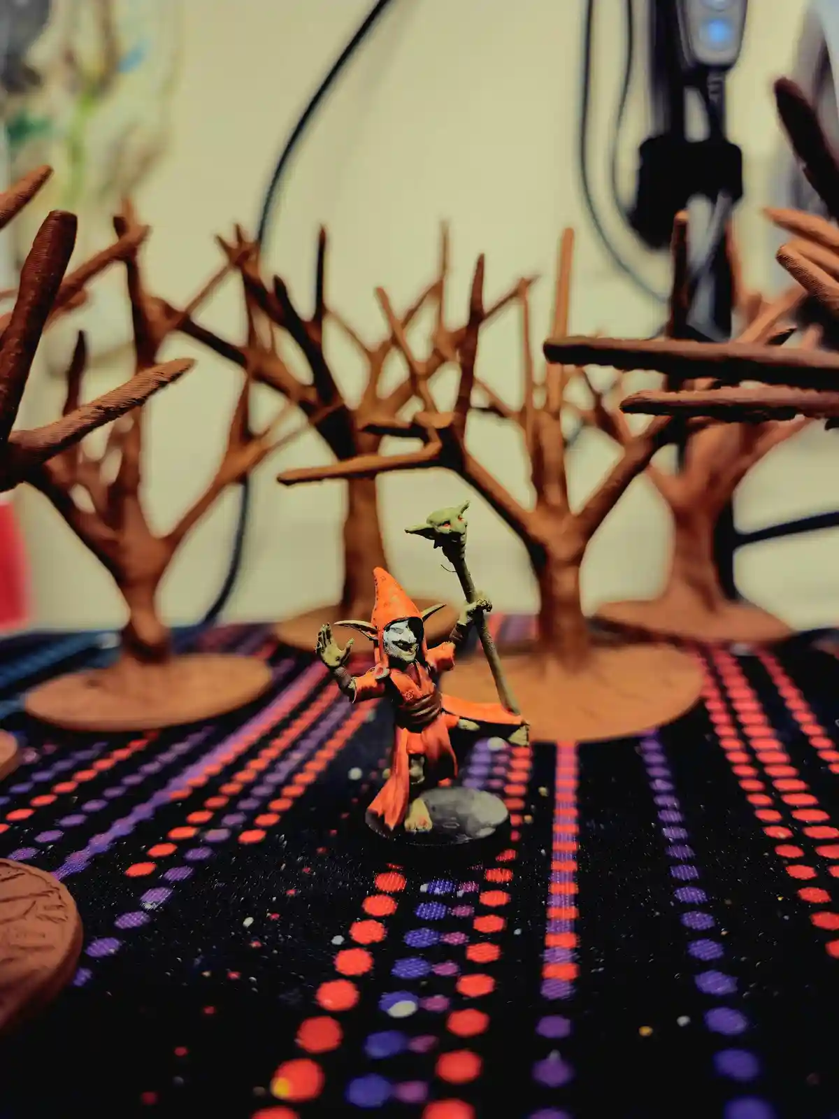 A painted miniature goblin wizard in an orange robe and pointed hat stands on a black, red, and purple patterned surface…