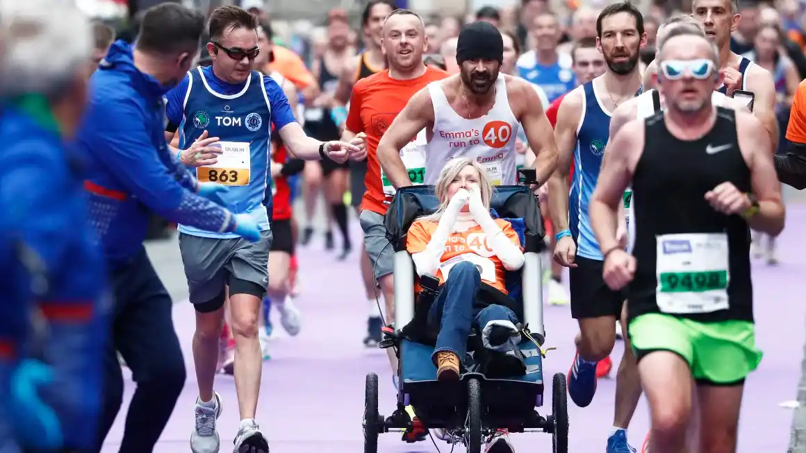 A woman in a wheelchair, wearing an orange shirt with 'Emma's run to 40' printed on it, is being pushed by a man in a white…