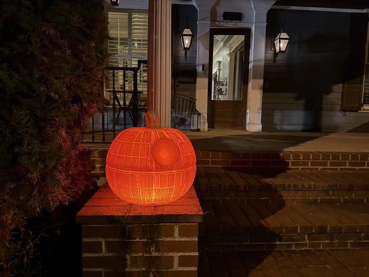 A glowing orange pumpkin, intricately carved to resemble the Death Star from Star Wars, sits on a brick pillar in front of a…