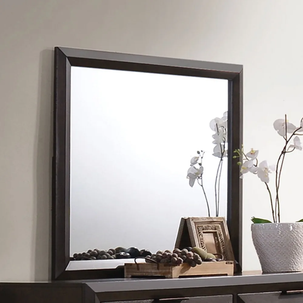 A dark-framed mirror sits on a dresser, reflecting a plain white wall while showing decorative items like white orchids, a…