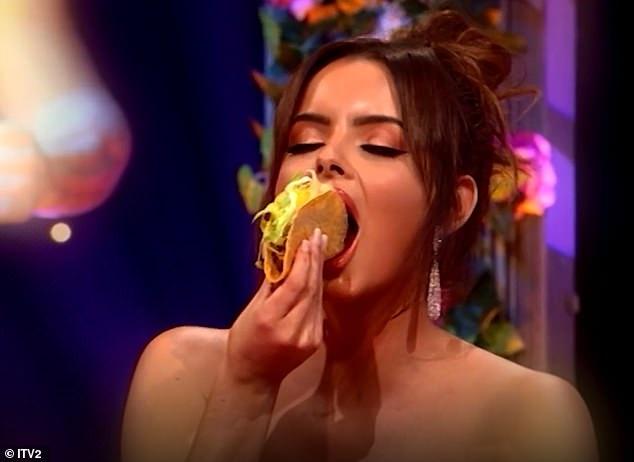 a woman with dark hair and long earrings is shown mid-bite, enthusiastically eating a large taco filled with lettuce and…