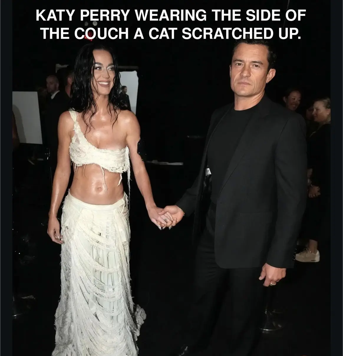 A photo of Katy Perry and Orlando Bloom holding hands at an event. Perry wears a distressed, shredded white gown that…