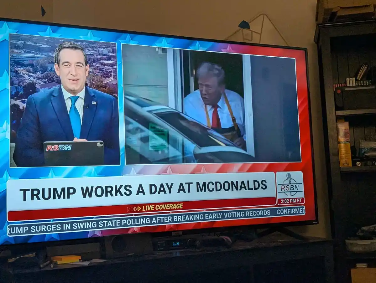 A television screen displays a news broadcast with a split screen: on the left, a news anchor in a suit and tie, and on the…