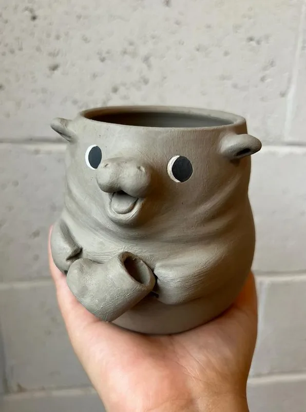 A hand holds a gray ceramic pot shaped like a cute, chubby animal with small ears, round black eyes, and a smiling mouth…