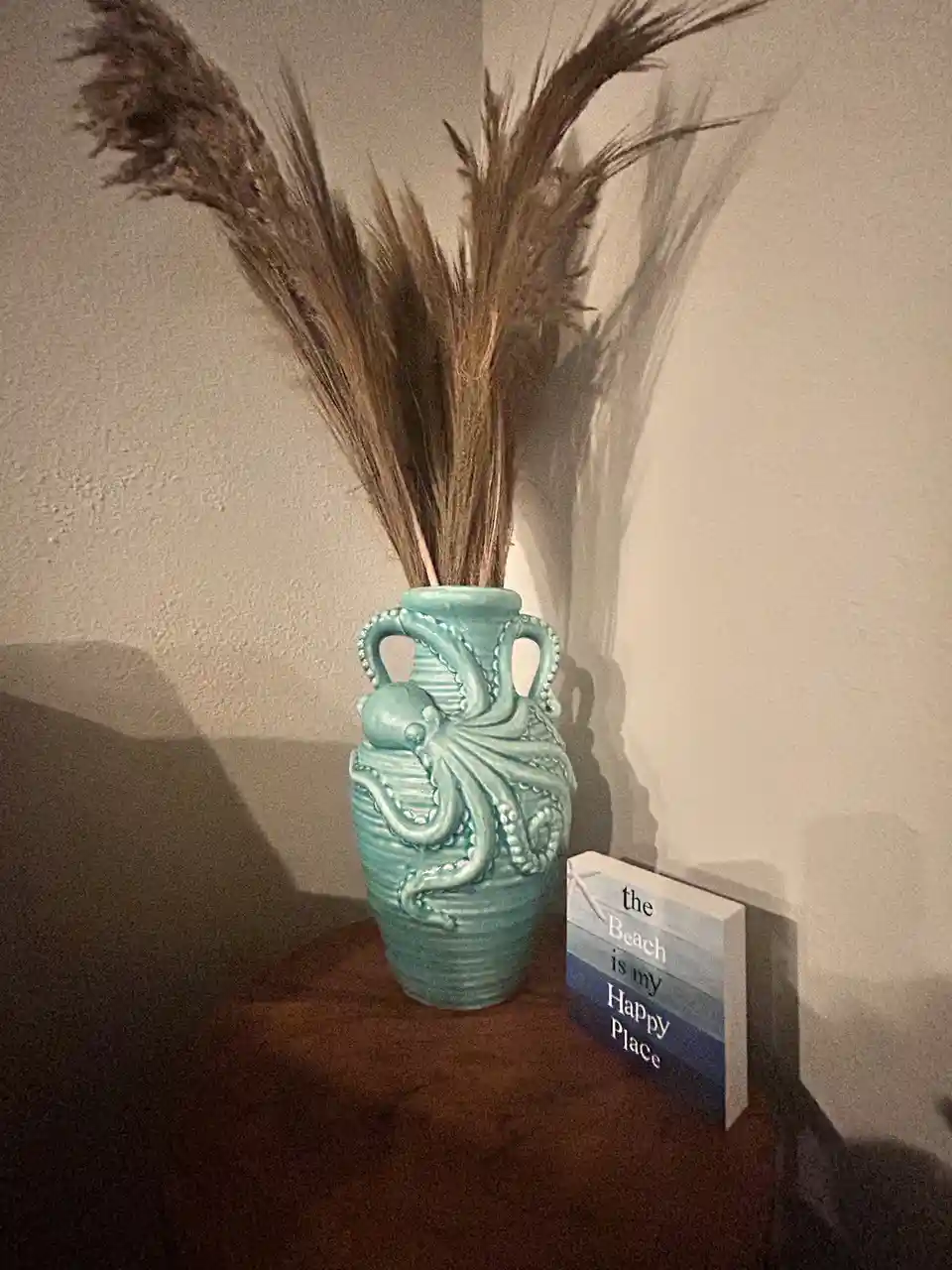 A teal ceramic vase shaped like an octopus holds dried pampas grass, sitting next to a small sign that reads 'the Beach is…