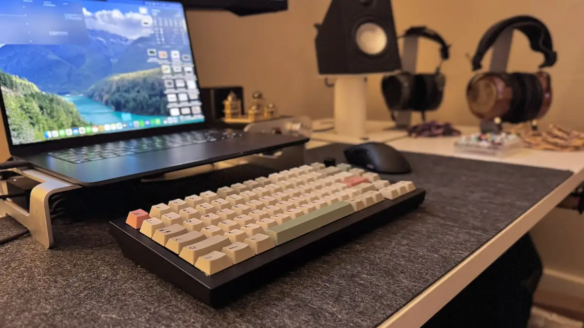 A close-up photo of a custom mechanical keyboard with beige and pastel-colored keys sits on a desk in front of a laptop on a…