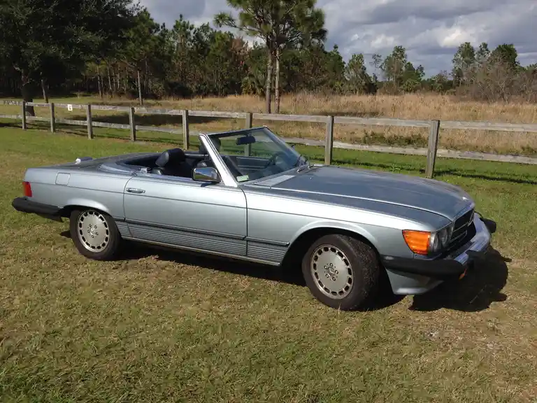 A silver Mercedes-Benz 380 SL convertible is parked on grass near a wooden fence with trees in the background. The car's top…