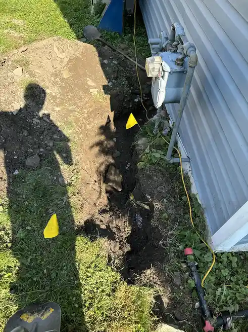 An outdoor photo shows a long, narrow trench dug in grass next to a building's corrugated metal siding, with a shovel nearby…