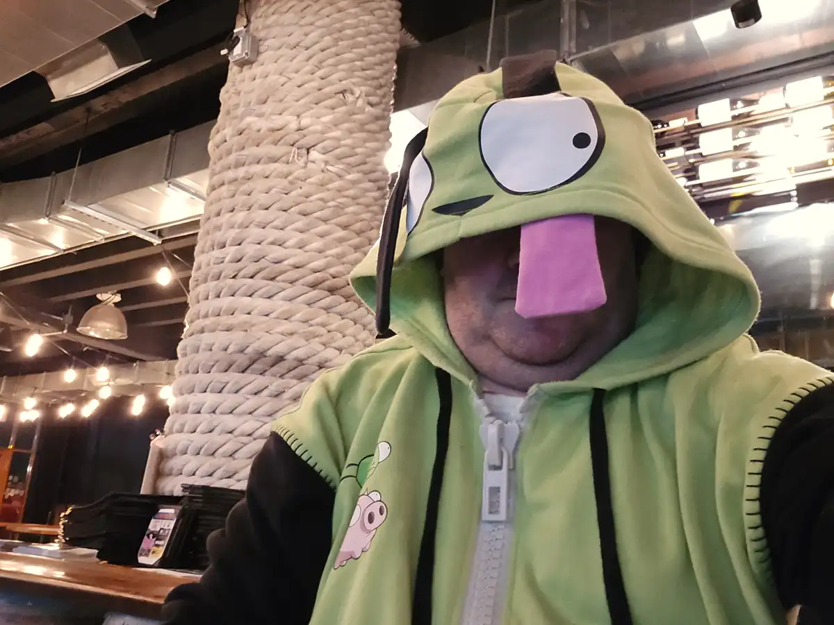 A person wearing a green, frog-like hooded costume with a large cartoon eye and a long pink tongue sticking out from the…