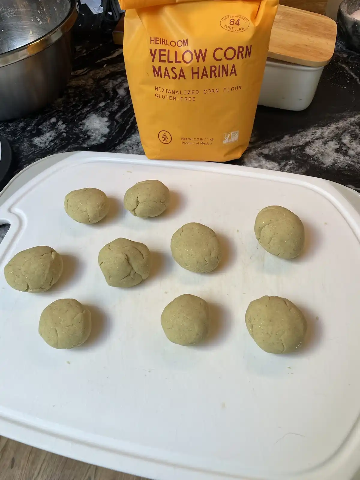 A white cutting board holds nine unevenly shaped, pale yellow dough balls, likely for tortillas, in front of a large yellow…
