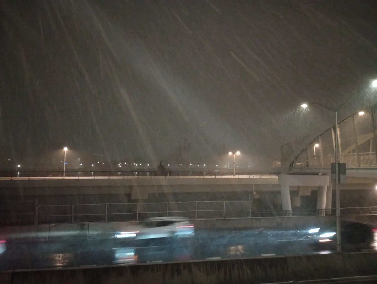 A nighttime photo taken from inside a vehicle shows heavy rain streaking vertically across the windshield, obscuring the…