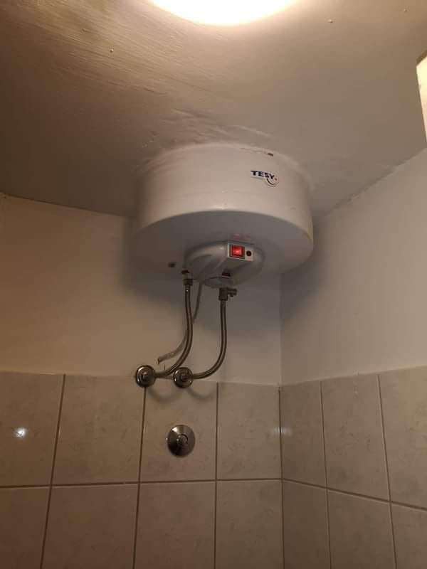 a white cylindrical water heater branded 'TESY' is visibly mounted upside down on a ceiling in a tiled bathroom corner, with…