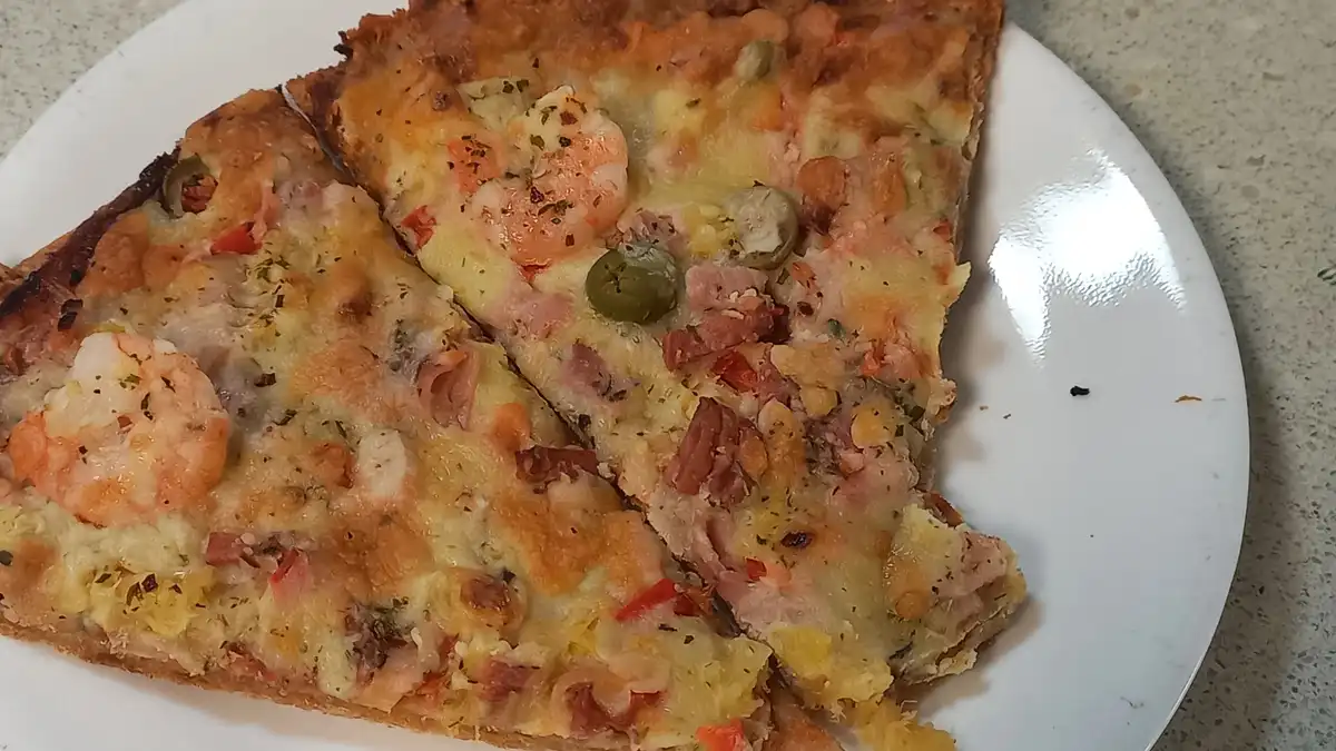 Two slices of a pizza with shrimp, ham, green olives, red peppers, and melted cheese are on a white plate. The crust is…