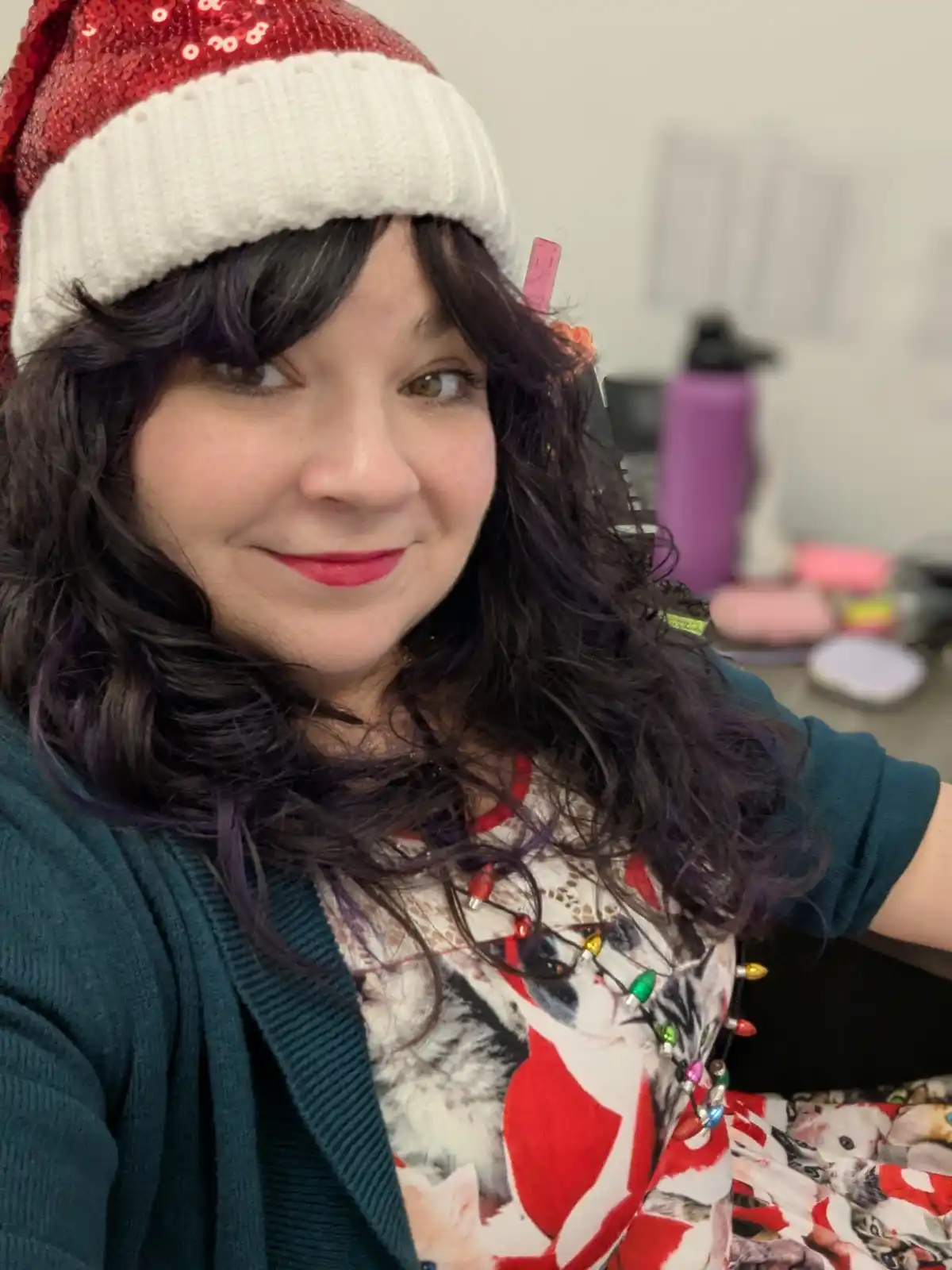 A woman with dark, wavy hair and red lipstick wears a red sequined Santa hat and a shirt with cats in Santa outfits, with…