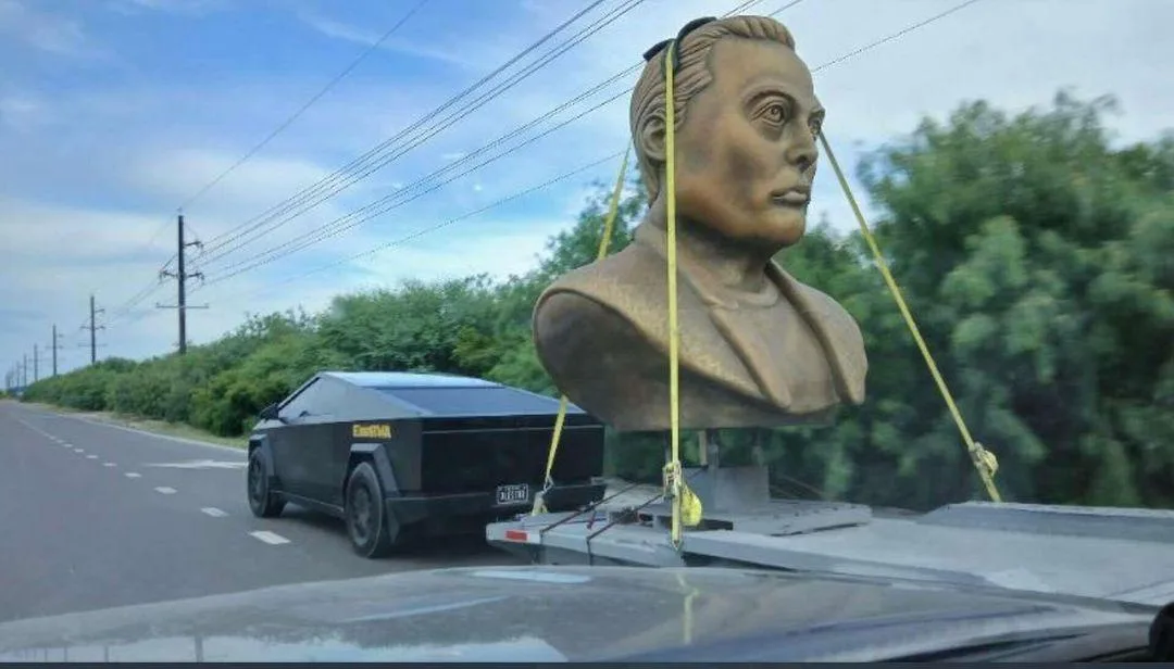 A black Tesla Cybertruck is towing a trailer with a large, bronze bust of Elon Musk strapped to the top with yellow straps…