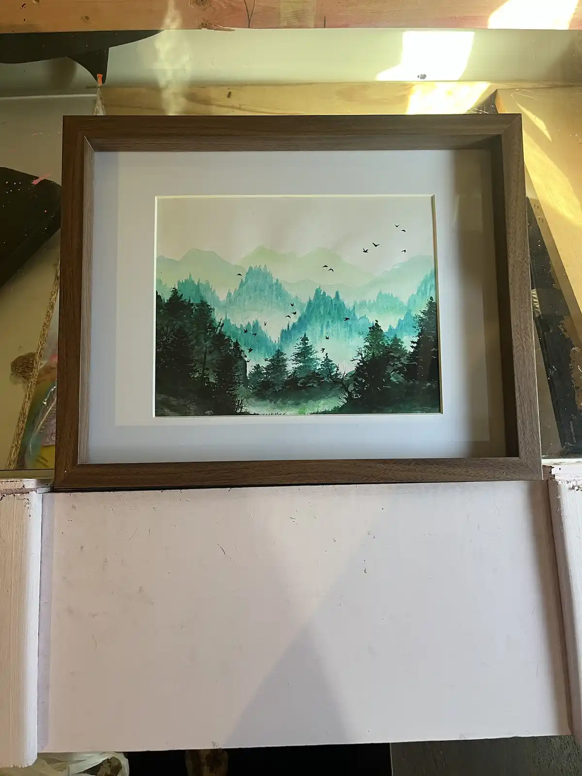 A framed watercolor painting of misty green mountains and forests with birds flying, displayed on a white surface. The…