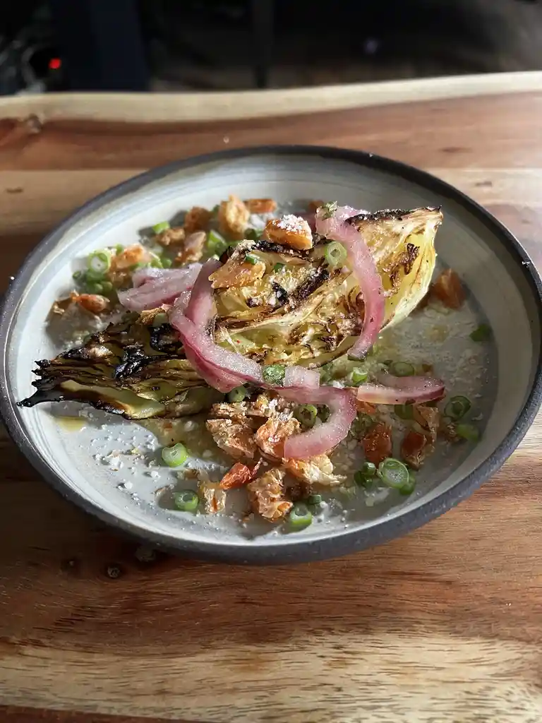 A close-up photo shows a ceramic bowl on a wooden table containing a dish with charred cabbage, pickled red onions, chopped…