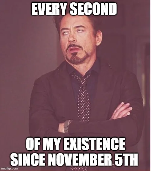 A man with a goatee and arms crossed, wearing a dark suit and tie, looks upward with a smug, slightly annoyed expression…