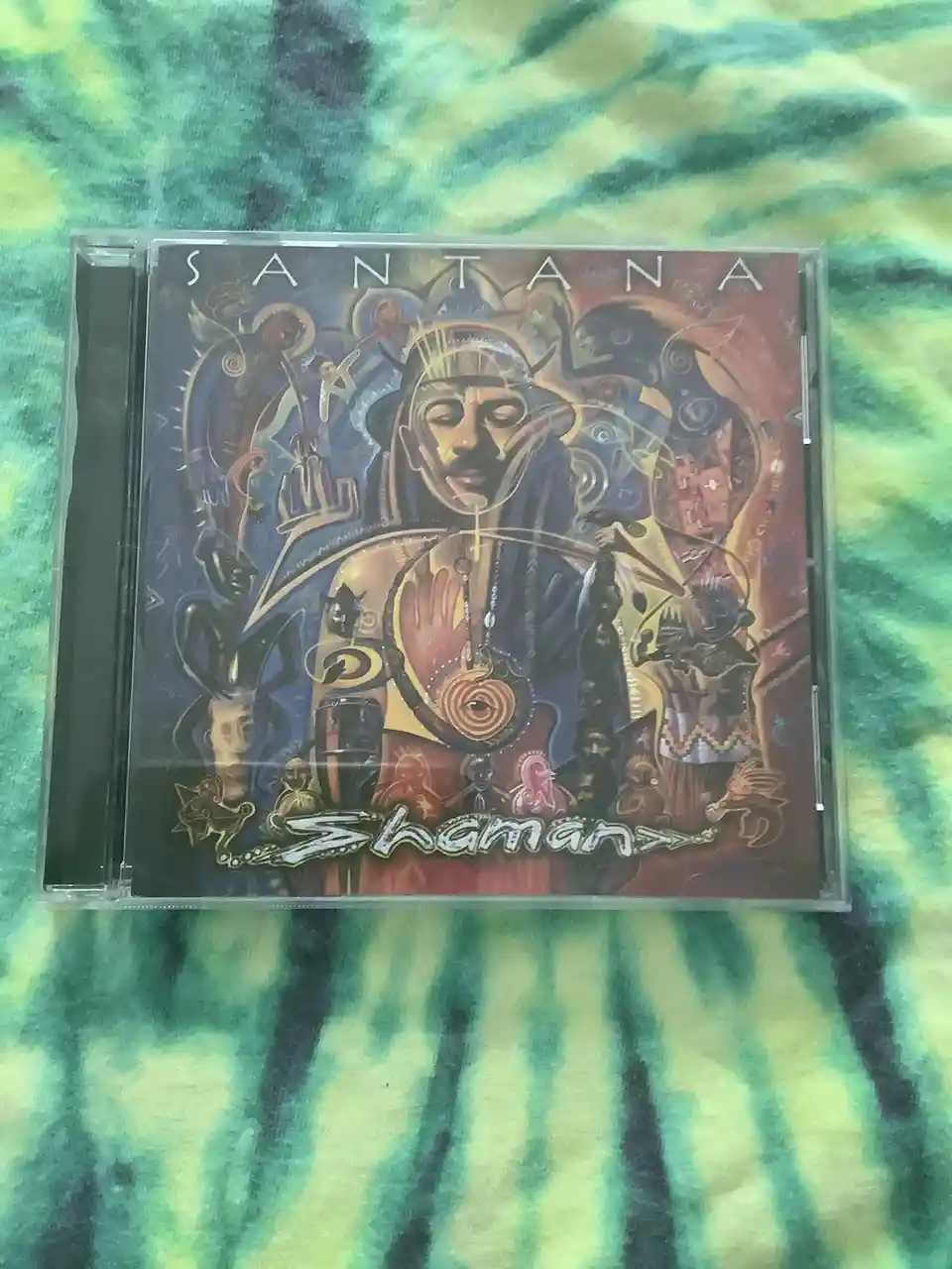 A CD case for Santana's 1999 album 'Shaman' rests on a green and yellow tie-dye fabric. The cover art features a central…