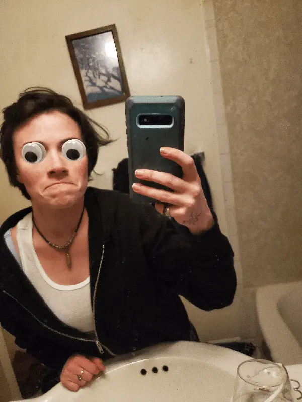 A person with large googly eyes stuck on their face takes three mirror selfies in a bathroom, making increasingly…