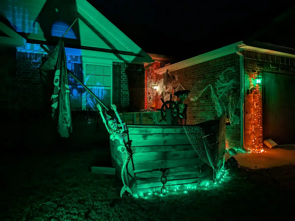 A nighttime photo of a house decorated for Halloween with a pirate theme. A wooden boat prop sits in the yard, illuminated…