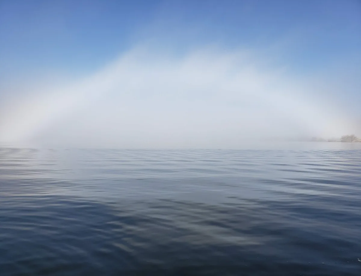 A calm, misty body of water under a clear blue sky, with a faint, white, arched mist hovering above the horizon, resembling…