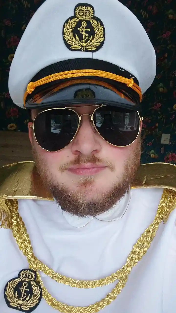 A man with a reddish beard and mustache wears a white naval officer's cap with a gold-embroidered anchor emblem, large…