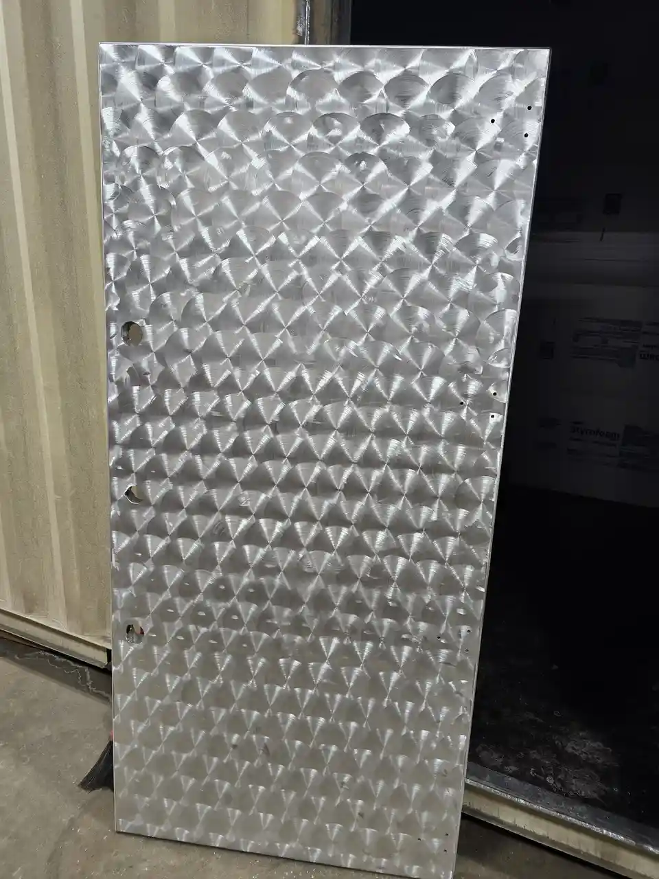 A large, vertically standing metal sheet with a textured, diamond-plate pattern and several holes punched into it, leaning…