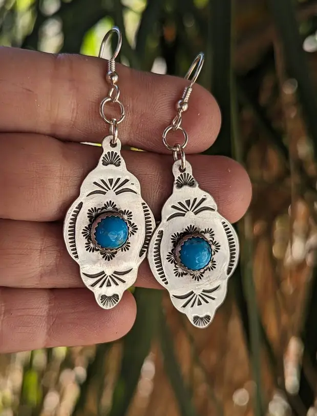 A hand holds a pair of silver earrings, each featuring a central turquoise stone surrounded by intricate stamped patterns…