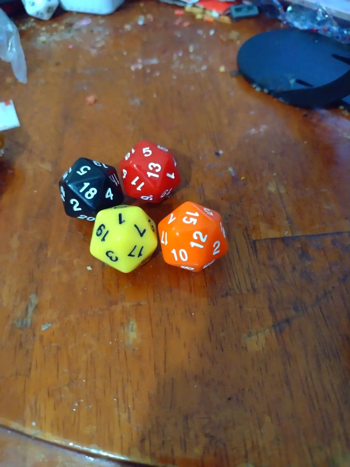 Four colorful twenty-sided dice (black, red, yellow, orange) rest on a wooden table, showing various numbers, with the…