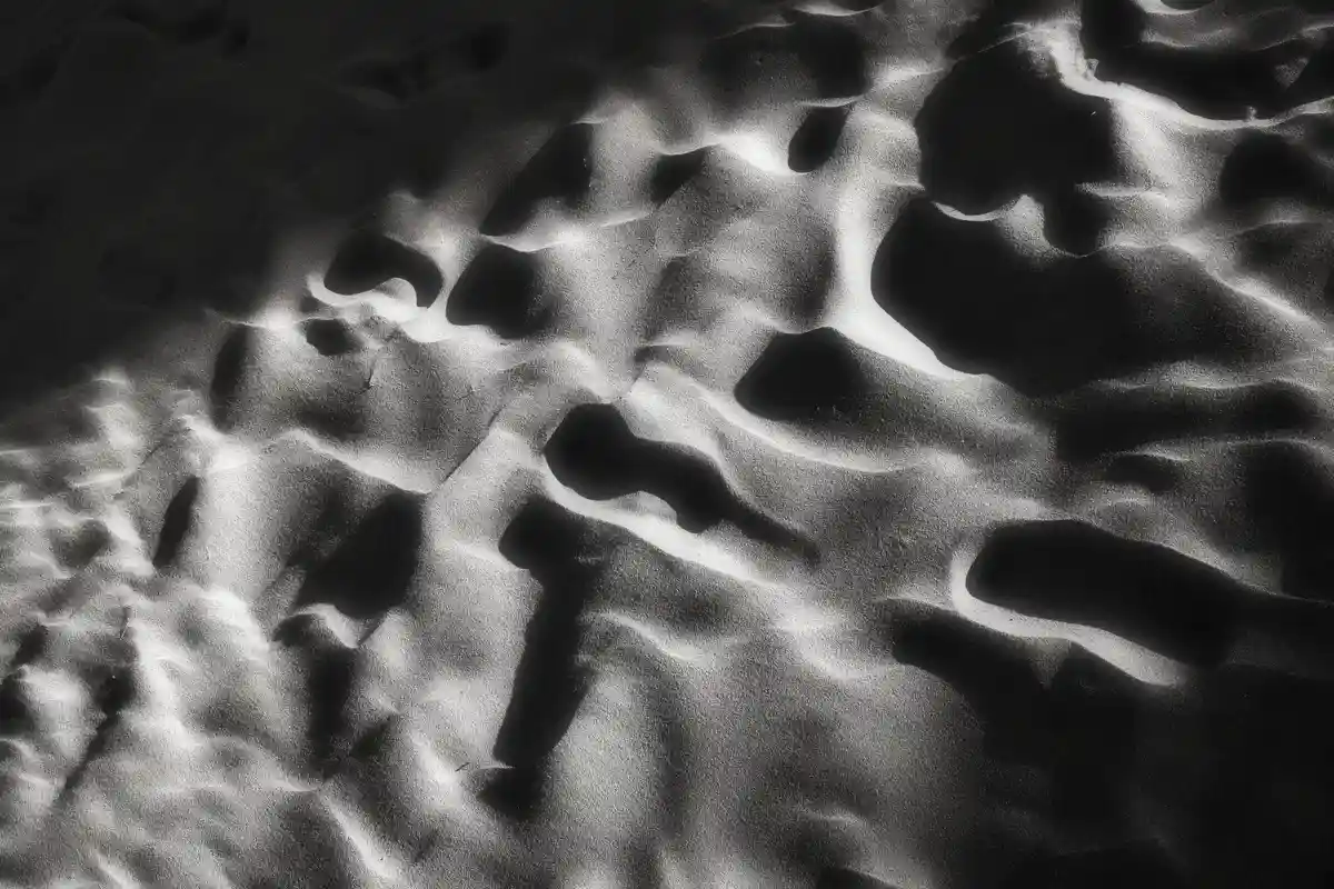 A black-and-white photograph of textured sand with dramatic lighting creating deep shadows and bright highlights. The…