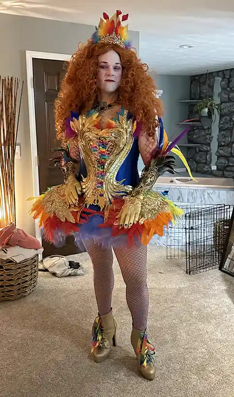 A person with curly red hair and heavy makeup stands in a living room wearing an elaborate, colorful costume with a rainbow…